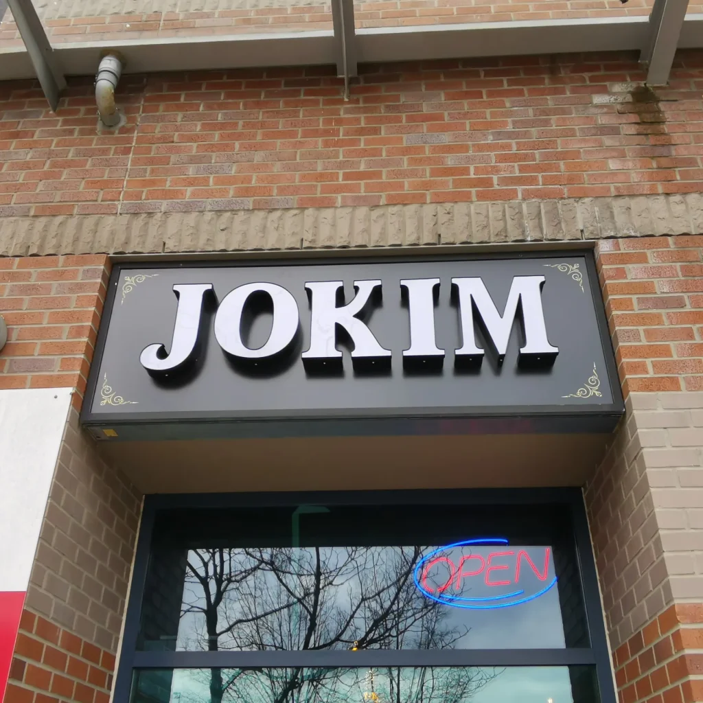 Channel letter sign with white face installed on a black sign box above a window of a restaurant.