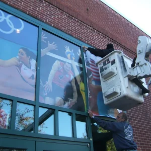 Two men installing window graphics on the exterior windows of a building, one is on a bucket truck.